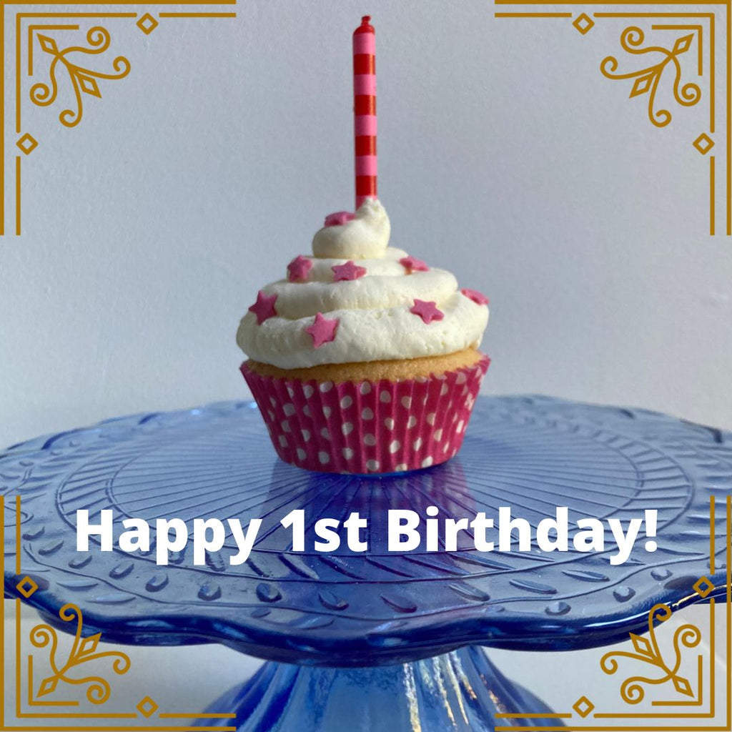 A picture of a celebration cupcake on a cake stand. It has one birthday candle on top. The caption reads Happy 1st Birthday.
