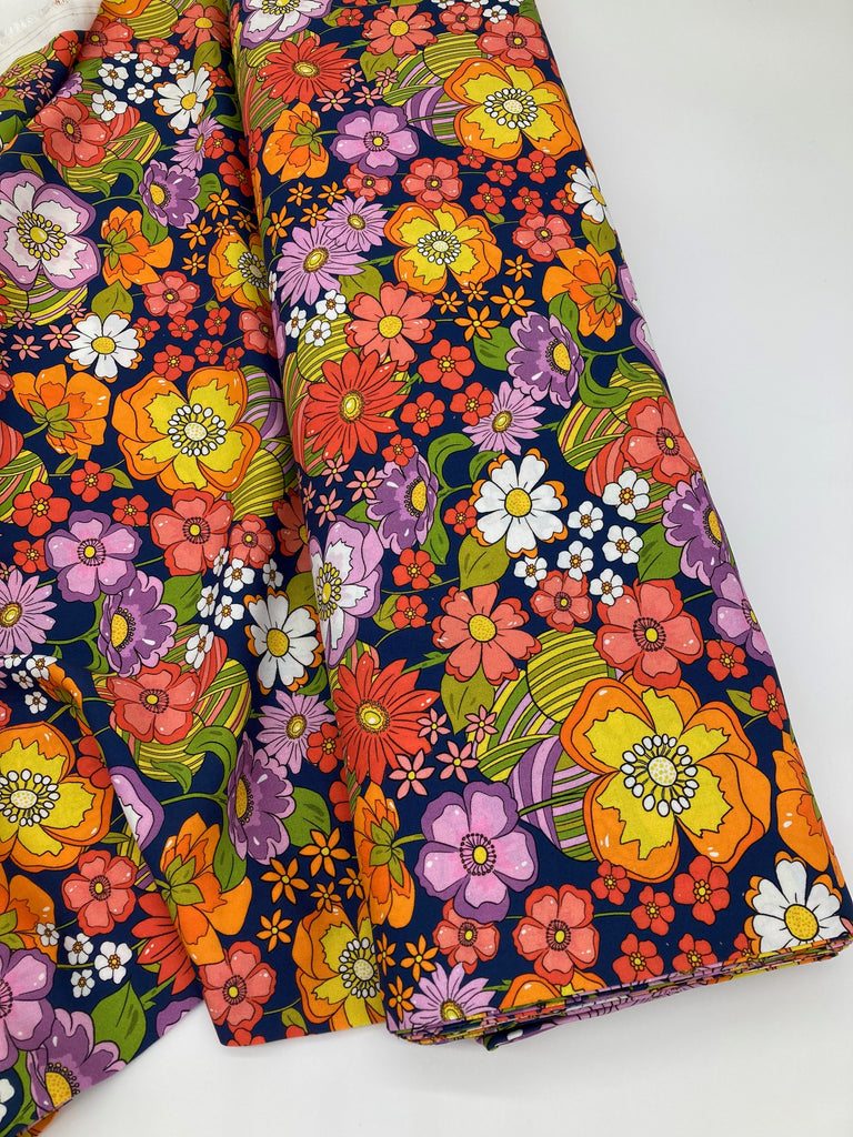 Fabric shown on a bolt. It is a retro floral design similar to 1970s prints, against a navy background.